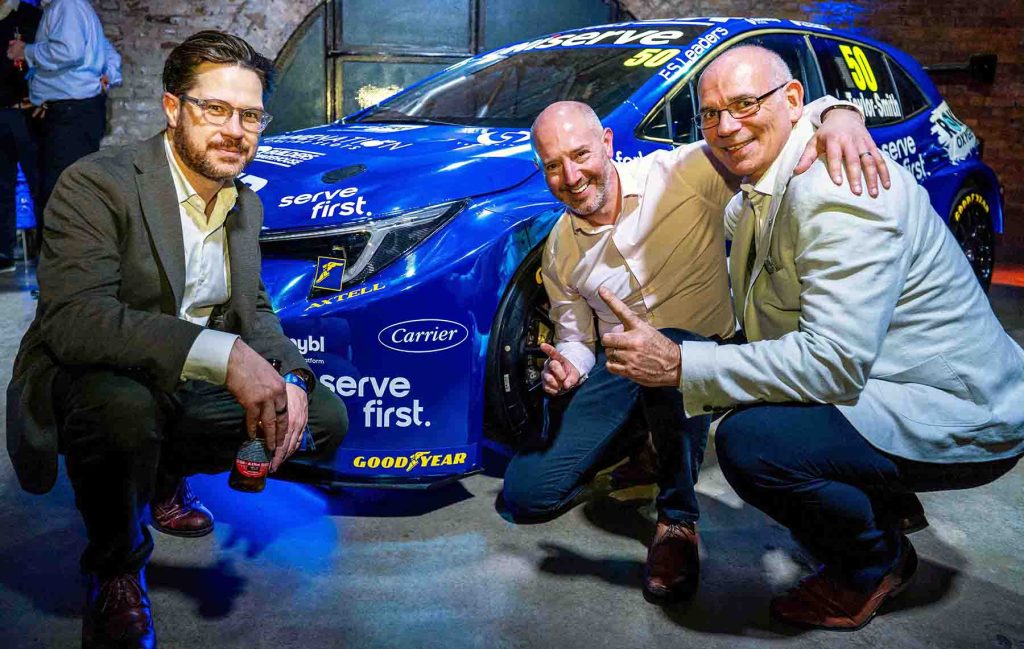 Three men with a blue race car, celebrating their partnership in racing.