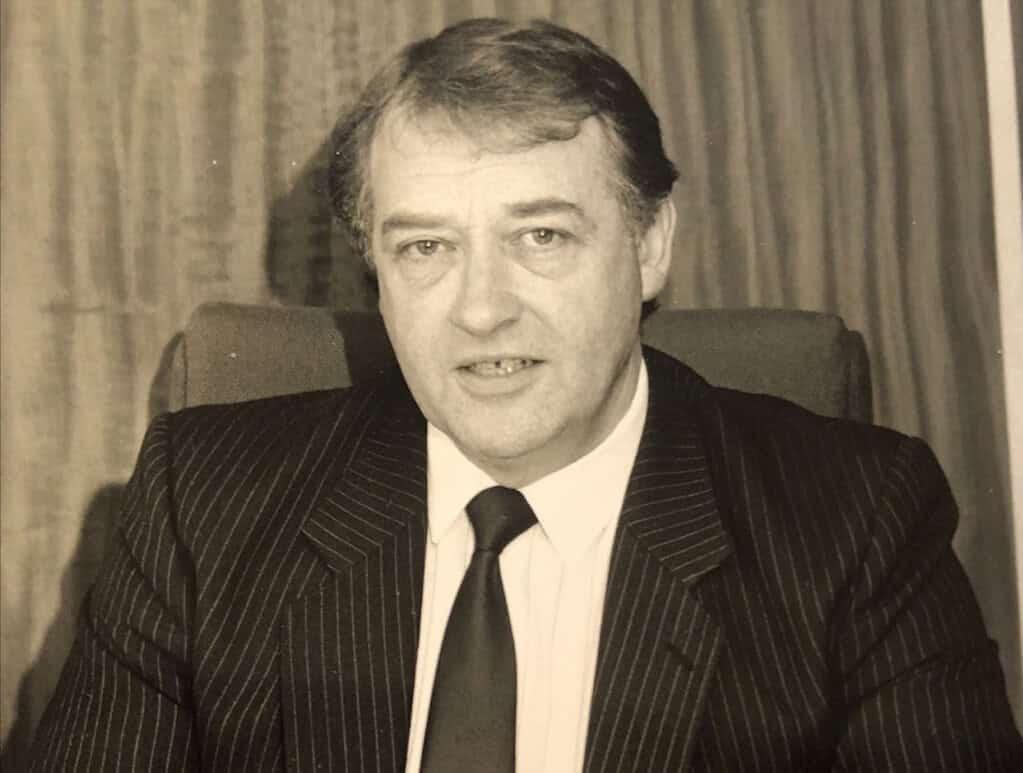 Business professional in formal attire, sitting at desk in an office setting.