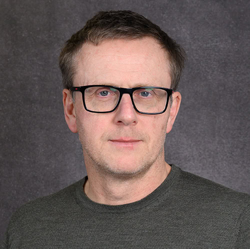 A man wearing glasses with a neutral expression, grey background.