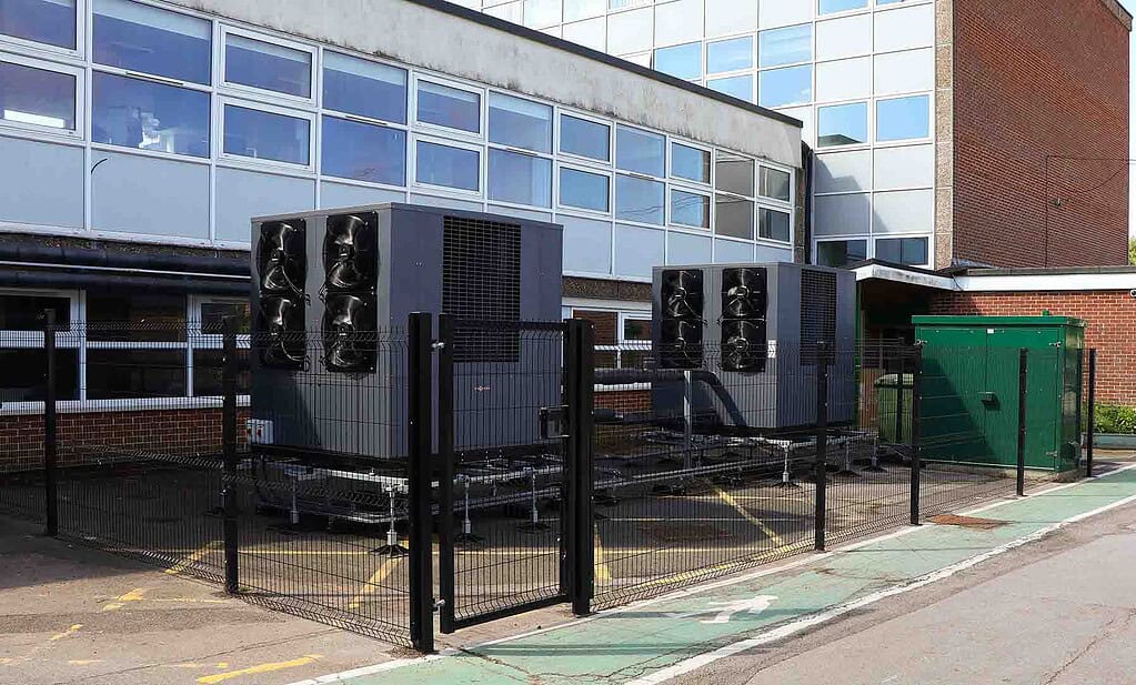 Air source heat pump units installed outdoors, enclosed by a black fence near a modern building.