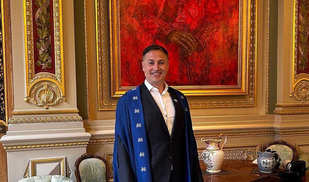 Man in blue academic gown smiling at a formal event with ornate decor.