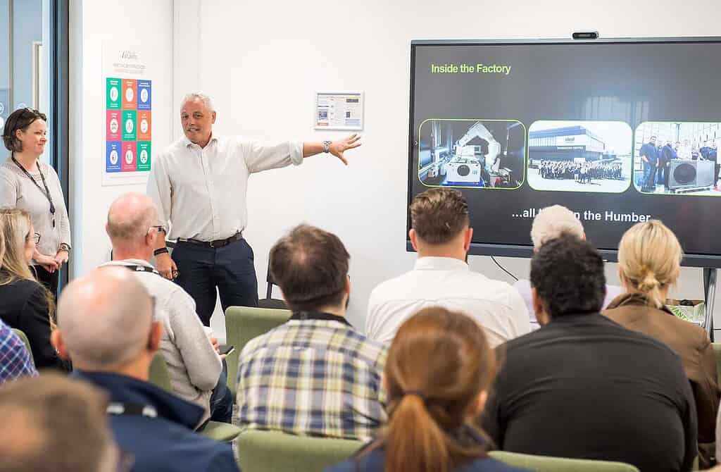 Heat pump presentation in a factory setting with a speaker and audience.