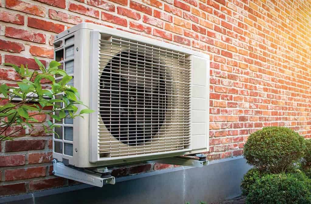 Outdoor heat pump unit installed on brick wall with greenery nearby.