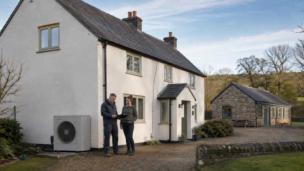 Two people discussing a heat pump outside a white house in the countryside.