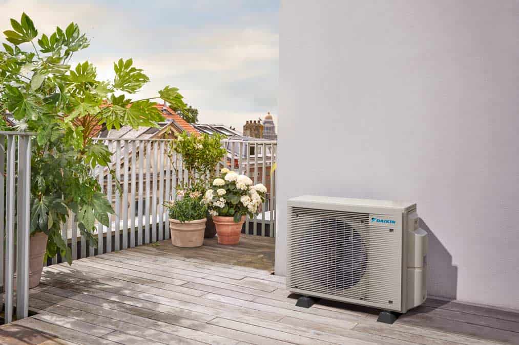 Outdoor heat pump on balcony with potted plants and city view.