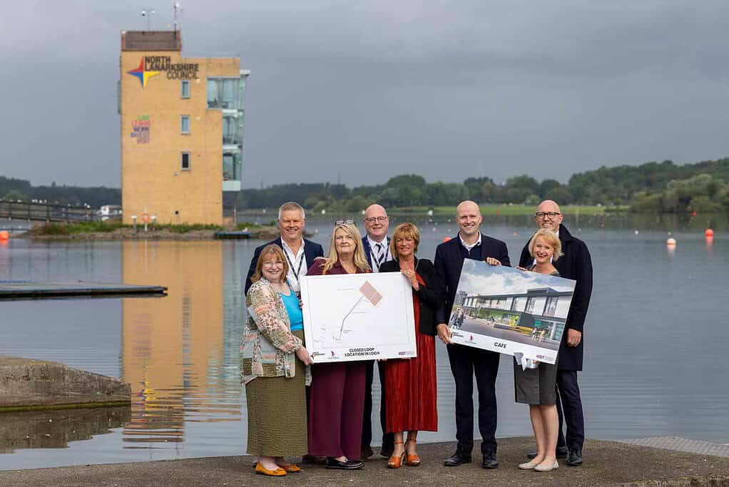 Group of people holding awards near Loch with building in background.