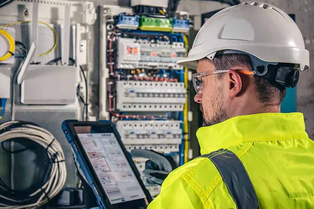Technician in safety gear inspecting electrical panel with tablet.
