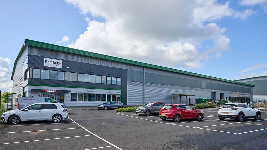 Large industrial warehouse with parking lot and cars under a partly cloudy sky.