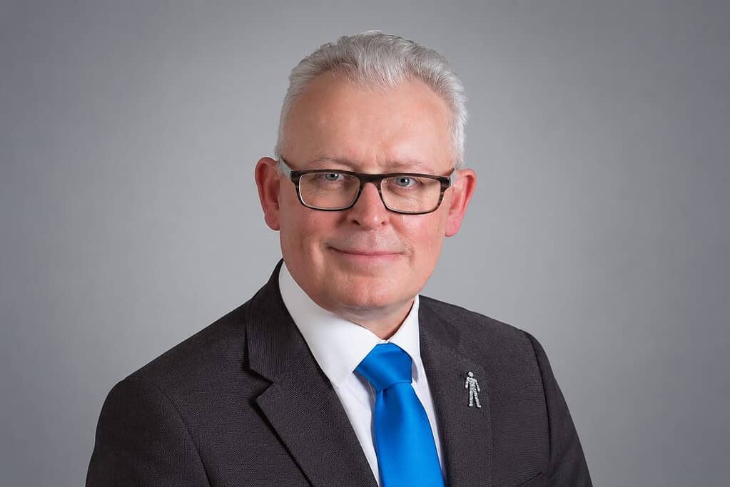 Professional portrait of Mark Hughes in a suit and tie with glasses.