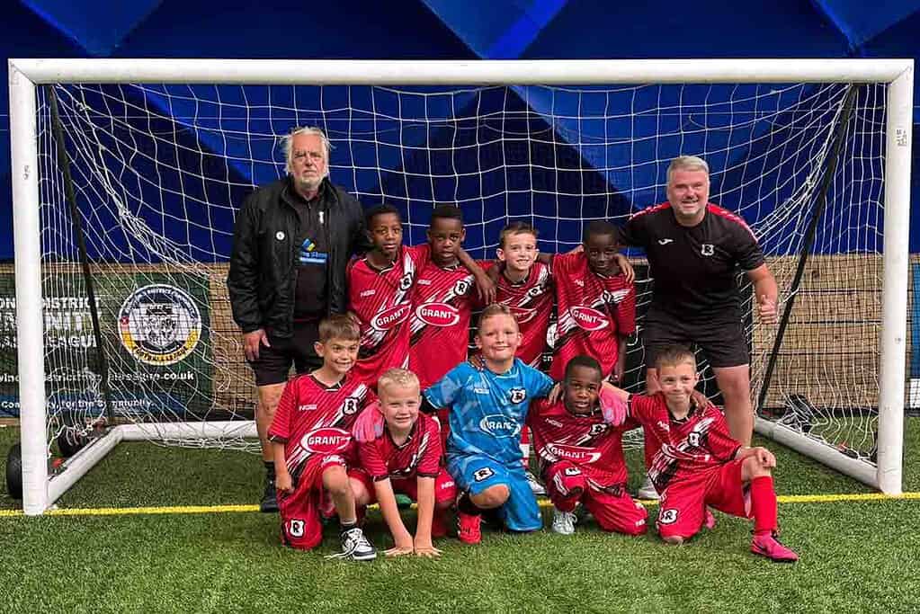 Youth soccer team with coaches posing in front of goalpost on artificial turf.