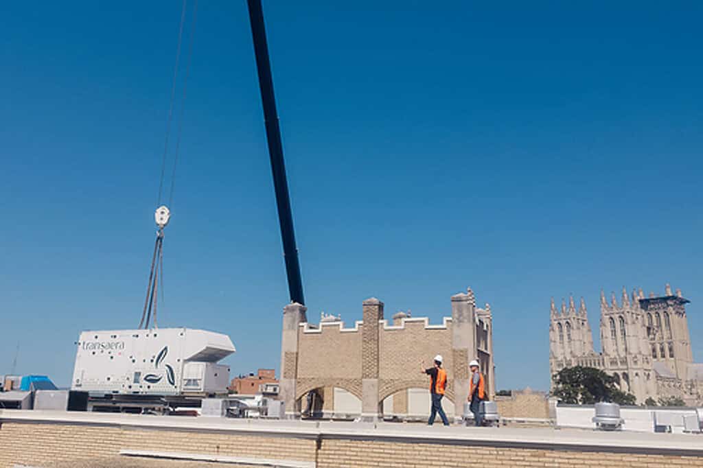 Large white transport container being lifted on a rooftop with workers nearby.